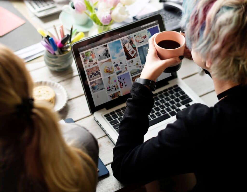 Two people at a desk, one with colorful hair, view a laptop displaying various images doing website maintenance. The person with colorful hair holds a coffee mug. The desk has flowers, a bowl, and stationery. Bright and creative atmosphere.