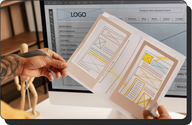 A person holds two wireframe sketches of a website design in front of a computer screen displaying website layout designs and UI elements. A wooden model and office furniture are seen in the background.