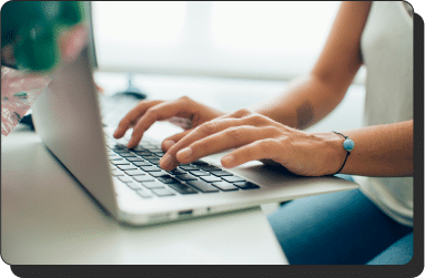 Close-up of a person typing on a laptop keyboard. The individual is wearing a white top and a bracelet with a blue bead. The background is slightly blurred, focusing on the hands and laptop.