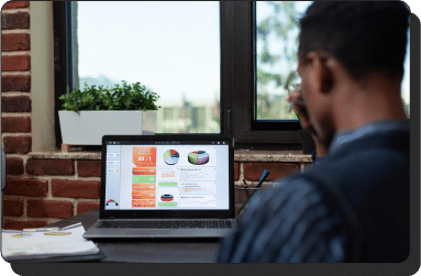 A person is sitting at a desk, looking at a laptop screen displaying colorful performance reporting charts and graphs. The desk is near a large window with a potted plant on the windowsill. There are papers and a pen next to the laptop. The background shows a brick wall.