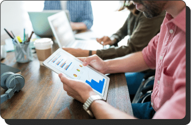 Marketers in a casual work setting are gathered around a wooden table with laptops and a tablet. The performance marketing professional in the foreground is holding a tablet displaying charts and graphs. Others are focused on their own devices, engaging in tasks or discussions.