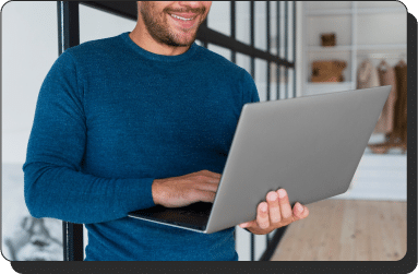 A performance marketer in a blue sweater stands indoors, smiling while using a laptop. He is leaning against a glass wall in a modern, stylish room with shelves in the background.