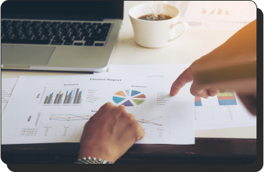 Two hands are pointing at printed financial reports on a desk. The reports display various performance marketing charts and graphs. A laptop and a cup of coffee are also on the desk, suggesting a work environment.