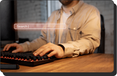 A person wearing a beige shirt is typing on a black keyboard at a desk. Above the keyboard, there is a digital search bar with the word "Search" inside it, glowing with a red tint. The background is a dimly lit room with brick walls.