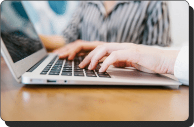 Close-up of a person's hands typing on a laptop keyboard. Another person is partially visible in the background, sitting at the same table, wearing a striped shirt. Both individuals are focused on the laptop screen. The setting appears to be a marketing workspace.