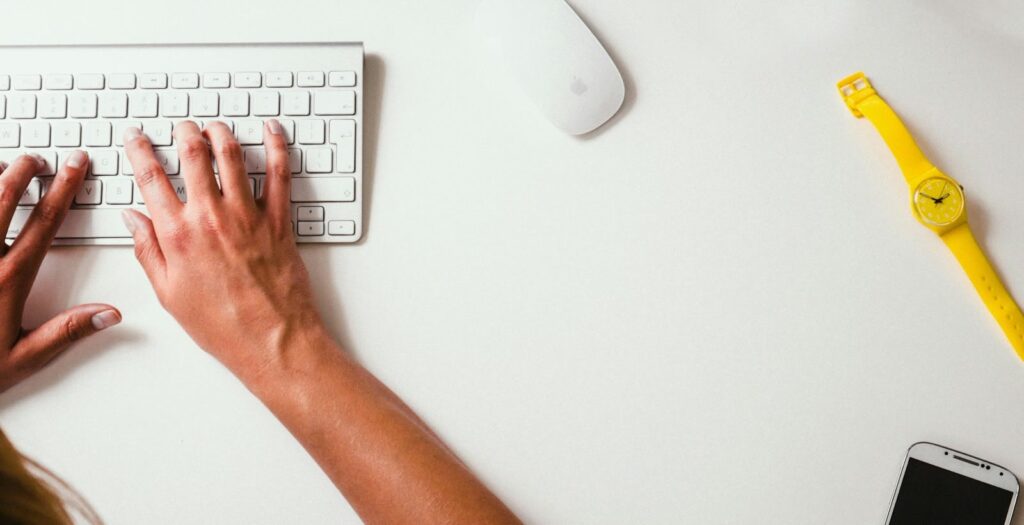 Hands typing on a white keyboard with a wireless mouse nearby on a clean, white desk doing regular website maintenance. A yellow wristwatch and a smartphone are placed on the right side of the desk.