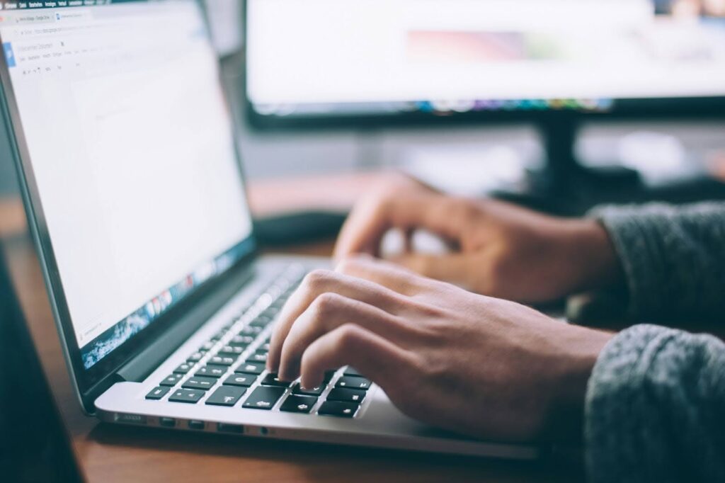 A person typing on a laptop keyboard, testing page speed, with a monitor in the background. The focus is on the hands, and the screen shows an open text document. The setting appears to be a workspace with soft lighting.