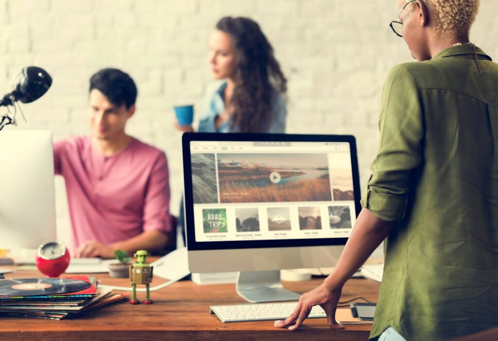 Three people in an office setting with computers and a tabletop with records discussing website maintenance. One person stands at a large monitor displaying a webpage, while the others, a seated individual and a person holding a cup, engage in discussion.