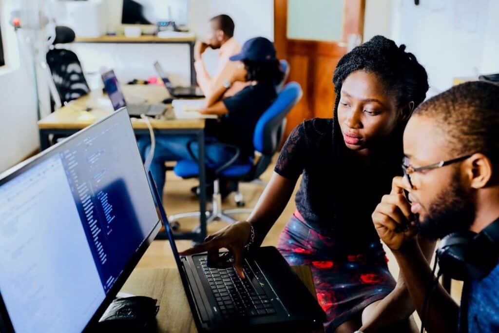 Two people collaborate at a desk with a computer, discussing page speed on the screen. The workspace is shared by others working in the background, suggesting an office or co-working environment.