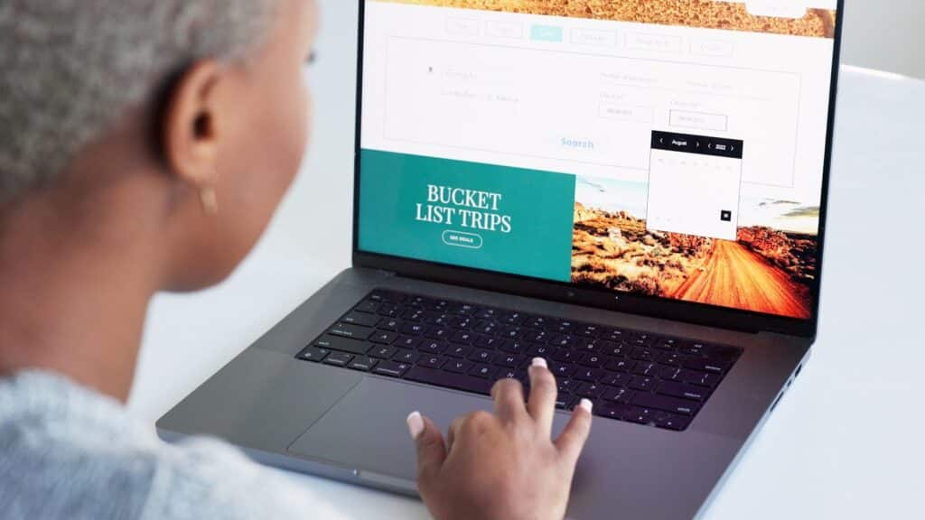A person using a laptop displaying a website with "Bucket List Trips" and an image of a dirt road. The person is typing on the keyboard while sitting at a white desk and doing website maintenance.