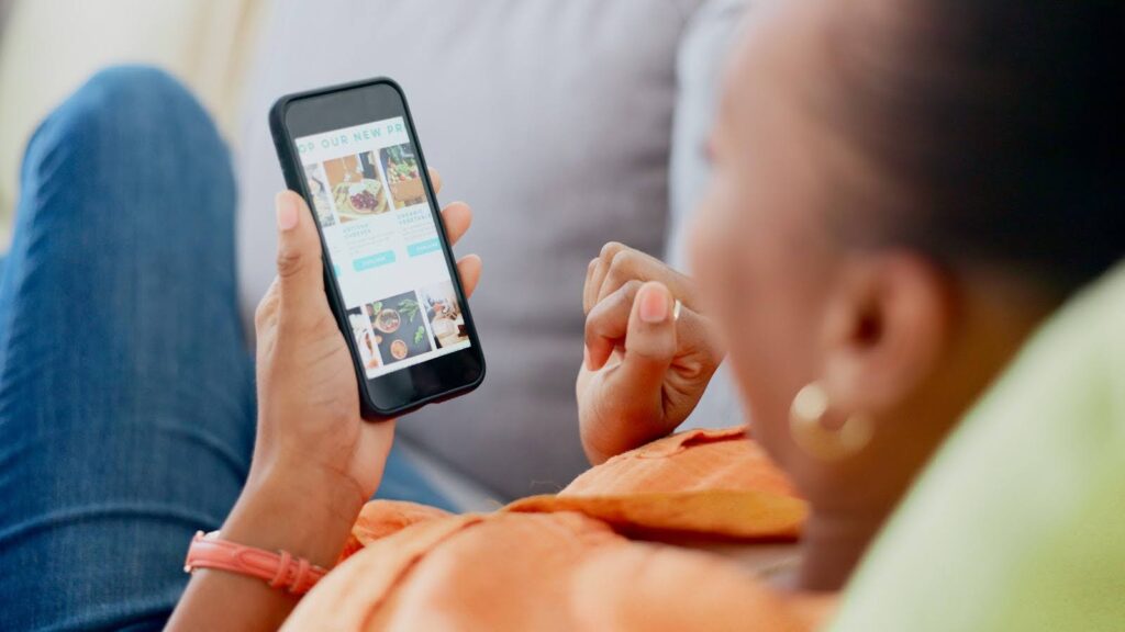 Person lying on a couch, holding a smartphone and testing page speed. The screen displays a food-related app with pictures and descriptions of various dishes. They are wearing an orange shirt and a red bracelet. The background is blurred.