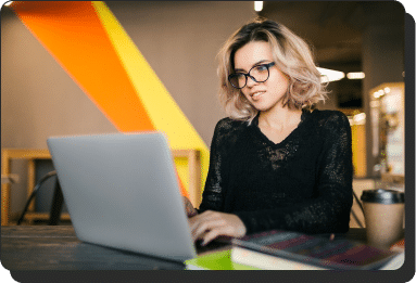 A person with glasses and shoulder-length hair is typing on a laptop researching lead generation services in a modern workspace. A colorful wall and a coffee cup are visible in the background.