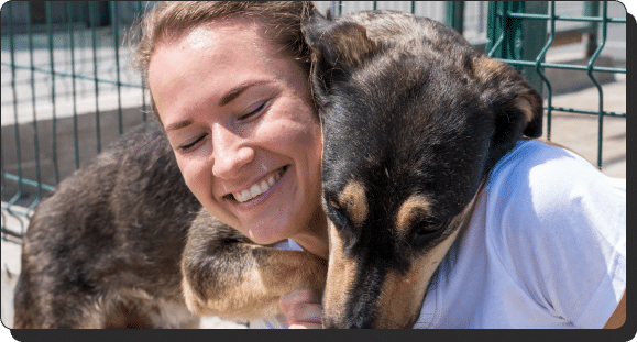 A smiling woman with closed eyes hugs a happy dog with a brown and black coat. The dog rests its head on the woman's shoulder. They are near a green fence, enjoying a sunny day.