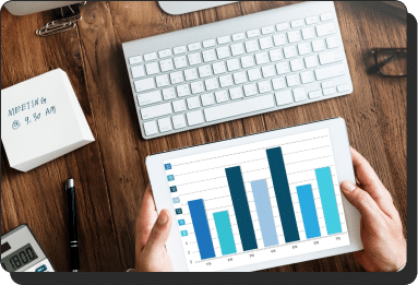 A person holds a tablet displaying a bar graph of business leads with varying shades of blue. Nearby are a keyboard, a notepad with "MEETING @ 9:00 AM," a stapler, and a calculator showing "1800," all on a wooden desk.