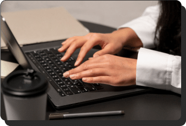 A person typing on a laptop keyboard at a desk. In the foreground, there is a coffee cup with a lid and a pen. A notebook is partially visible in the background. The person is wearing a white shirt.
