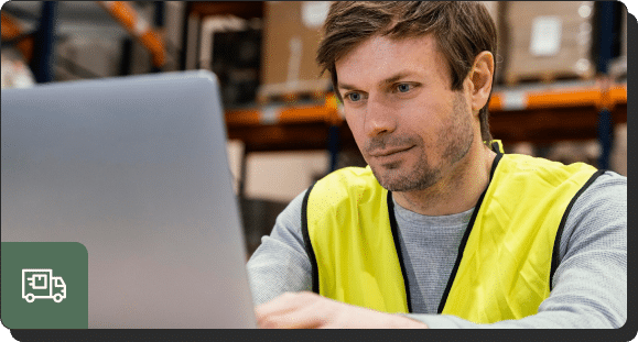 A man wearing a yellow safety vest is focused on looking up lead generation strategies on a laptop in a warehouse setting. Shelves with boxes are visible in the background.