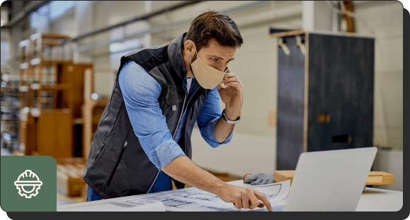 A man wearing a mask and a vest is speaking on a phone while working at a desk. He is looking at a laptop screen and pointing at a sheet of paper with architectural plans in a workshop or industrial setting.