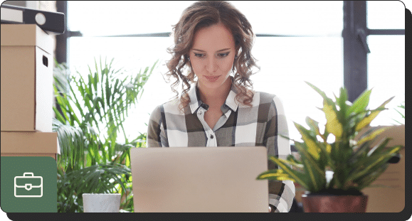 A woman with curly hair in a plaid shirt works on a laptop checking her marketing analytics in a bright room filled with plants and boxes. She appears focused, and there is a suitcase icon in the lower left corner of the image.