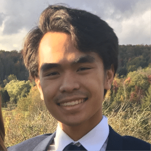 A smiling person with dark hair in a suit and tie stands outdoors. The background features a scenic landscape with trees and a cloudy sky.