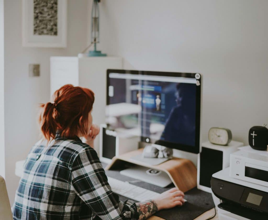 A person with red hair in a plaid shirt sits at a desk, using a computer with a large monitor to manage PPC advertising in a home office setting. Various office items and electronics are on the desk.