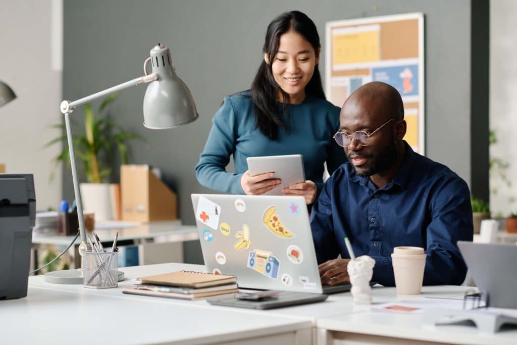 Two coworkers in an office collaborate on PPC advertising; one is seated at a laptop with colorful stickers, while the other stands nearby holding a tablet. Both appear engaged and focused on outsourcing PPC tasks.
