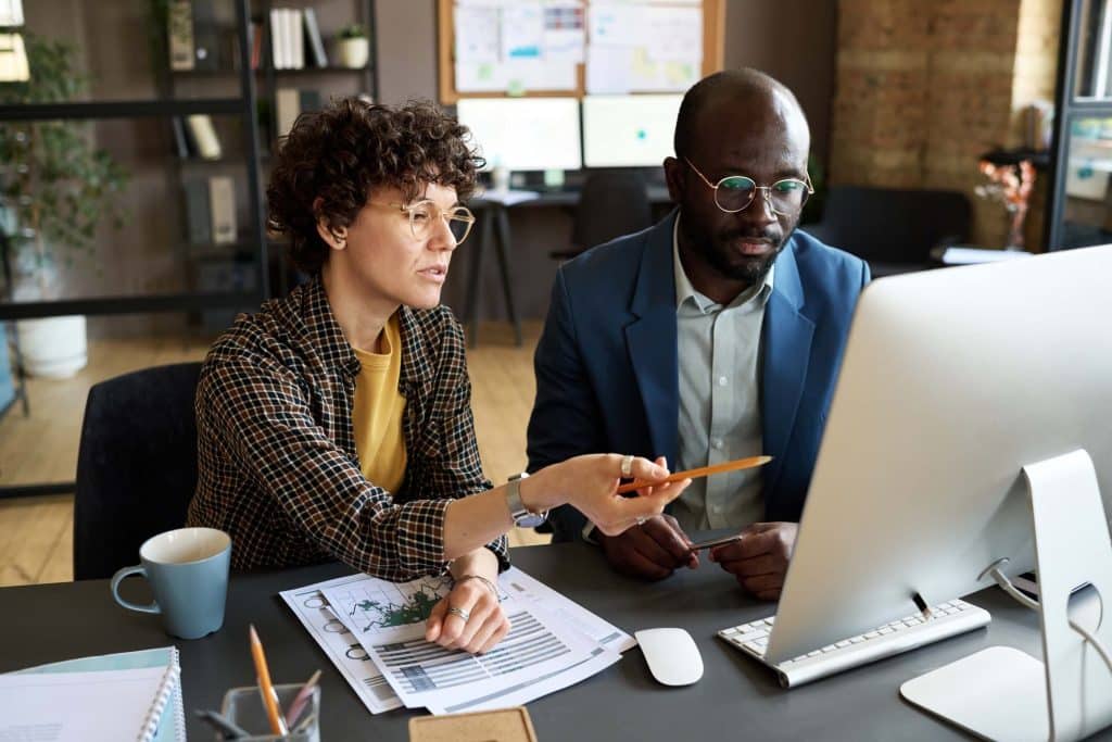 Two coworkers sit at a desk, discussing PPC outsourcing strategies as they review data on a computer screen. One points at the screen with a pencil while the other listens attentively. Documents, a notebook, and a coffee mug are on the desk.