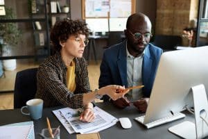 Two coworkers sit at a desk, discussing PPC outsourcing strategies as they review data on a computer screen. One points at the screen with a pencil while the other listens attentively. Documents, a notebook, and a coffee mug are on the desk.