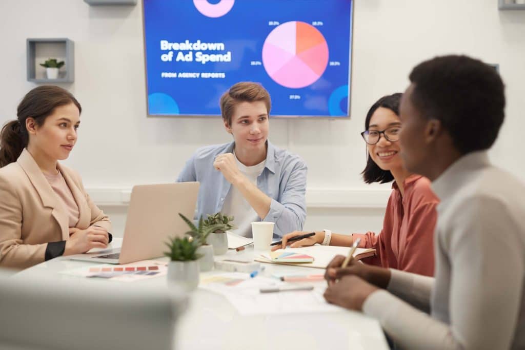 Four people sit around a table with laptops and papers, engaged in discussion about ppc outsourcing. A screen behind them displays a pie chart titled “Breakdown of Ad Spend” in a modern office meeting room.