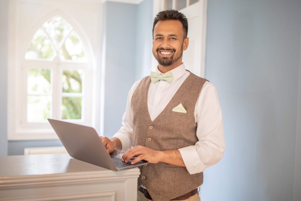 Owning a Wedding Venue: What It Actually Takes to Build a Booked-Out Calendar 2 A venue owner in a vest, bow tie, and dress shirt stands indoors, smiling while using a laptop on a white counter. A large window with arched panes lets in natural light behind him at the elegant wedding venue.