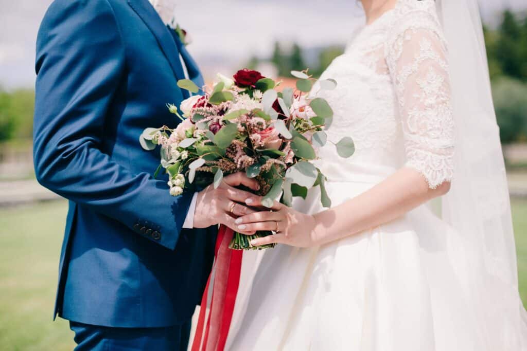 Owning a Wedding Venue: What It Actually Takes to Build a Booked-Out Calendar 1 A bride in a white dress and a groom in a blue suit stand close together outdoors at their charming wedding venue, holding a bouquet of red, pink, and white blooms, while the bride’s hand displays her wedding ring.