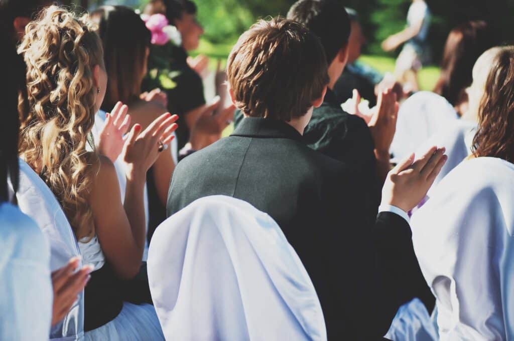 Owning a Wedding Venue: What It Actually Takes to Build a Booked-Out Calendar 3 Guests dressed formally, seated outdoors at a wedding venue, face away from the camera and applaud. Sunlight highlights their hair and clothing, white chair covers and lush greenery set the scene—perfect inspiration for anyone owning a wedding venue.