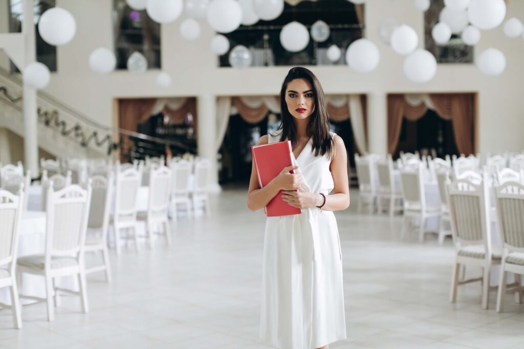 Owning a Wedding Venue: What It Actually Takes to Build a Booked-Out Calendar 4 A woman in a white dress stands in an elegant wedding venue, holding a red folder. White chairs and round hanging decorations fill the bright, spacious room, reflecting the care of a dedicated venue owner.